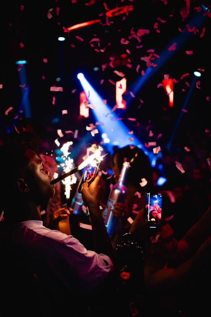 Homme filme étincelles dans une discothèque animée. Confettis roses tombent, lumières bleues et rouges. Ambiance festive et énergique.