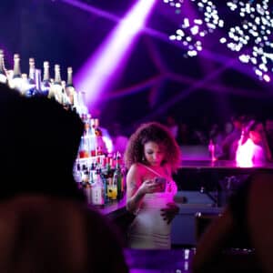 Femme aux cheveux bouclés dans une boîte de nuit vibrante, regardant son téléphone au bar éclairé par des lumières violettes et des bouteilles.