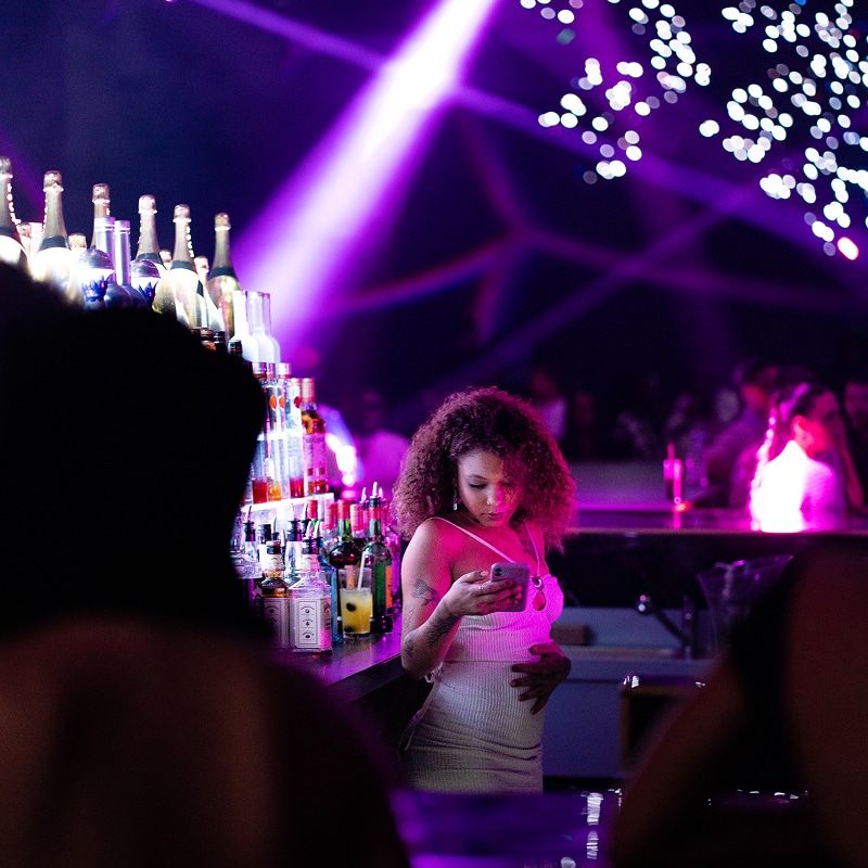 Femme aux cheveux bouclés dans une boîte de nuit vibrante, regardant son téléphone au bar éclairé par des lumières violettes et des bouteilles.