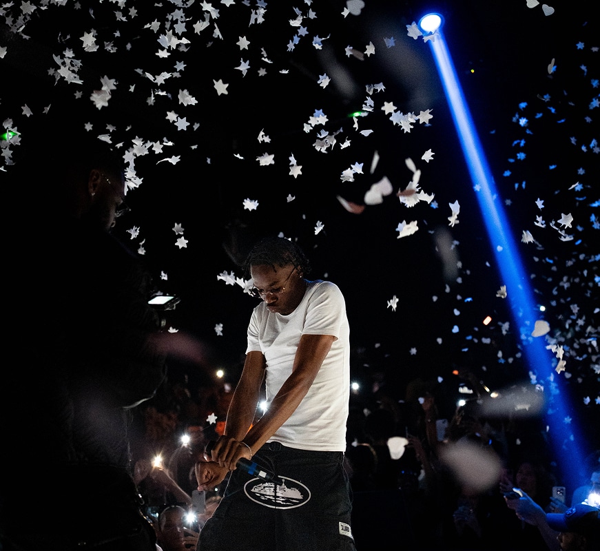Artiste sur scène au micro, devant un public éclairé par des téléphones, sous un faisceau lumineux bleu et des confettis en forme d'étoiles.