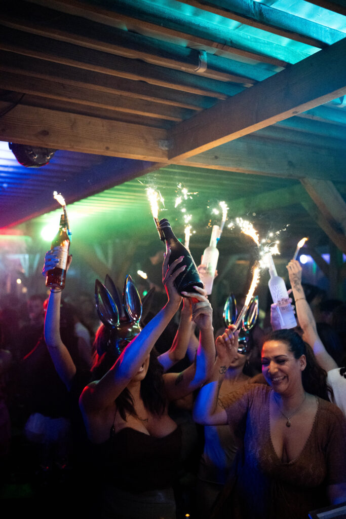 Fête animée en club: personnes lèvent des bouteilles avec feux de Bengale scintillants. Une femme souriante danse, d'autres portent des oreilles de lapin.