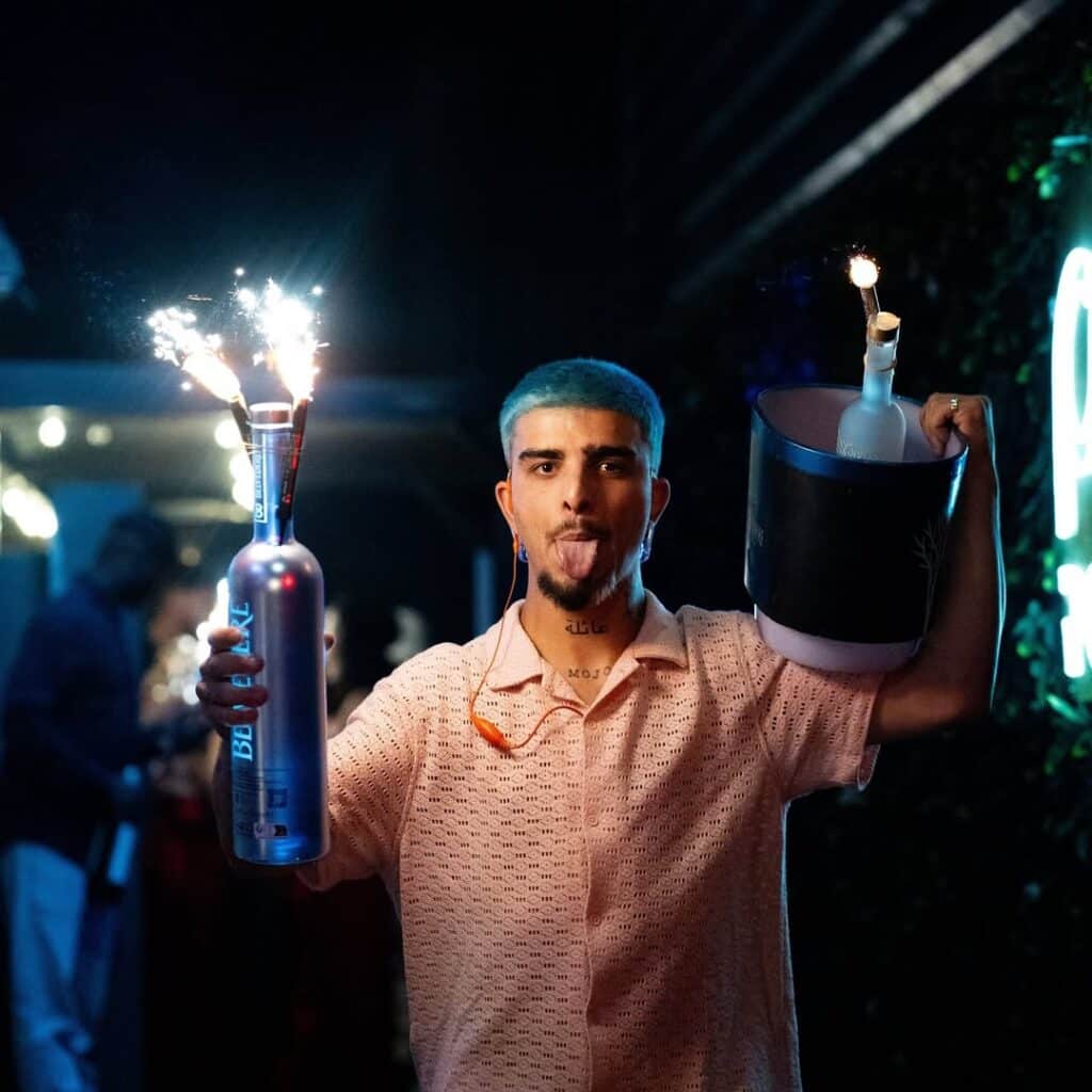 Homme aux cheveux bleus sortant la langue, tenant une bouteille de Belvedere avec feux d'artifice et un seau à glace. Tatouages 'MOJO' et 'عائلة' visibles.