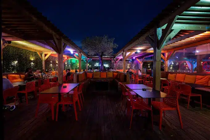 Terrasse lounge Le Miraye de nuit : pergolas en bois, lumières colorées, banquettes orange, tables, clients détendus et arbre central.