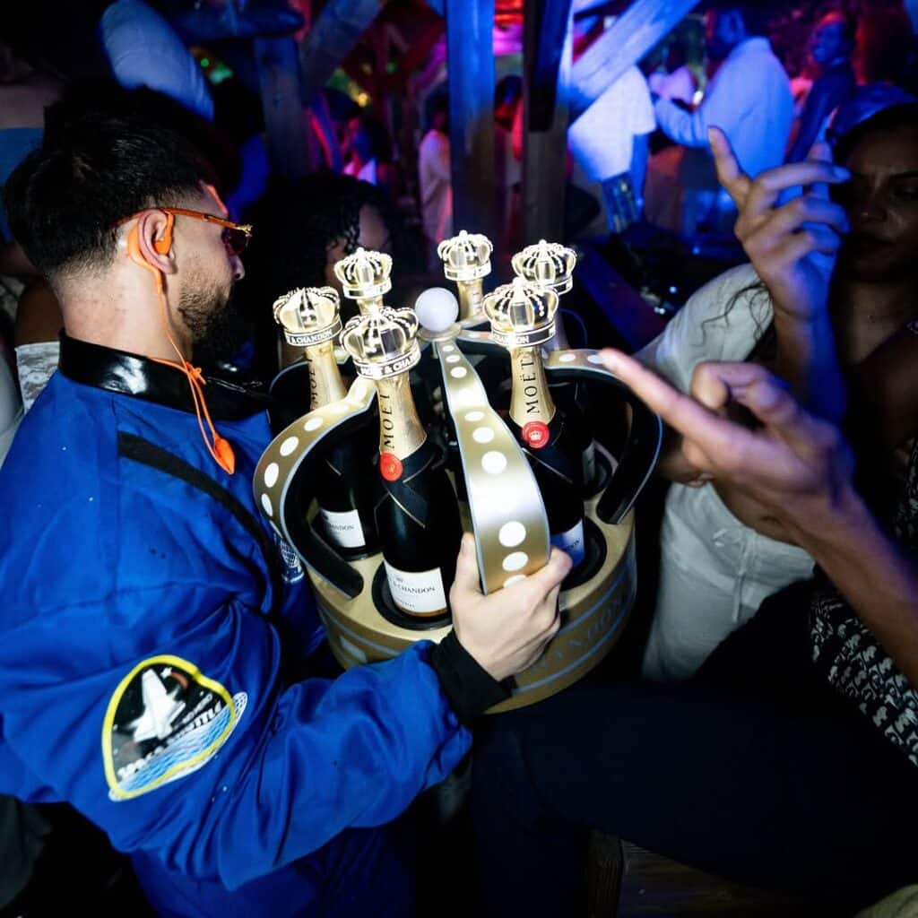 Homme en costume d'astronaute bleu tient un seau à champagne Moët & Chandon lumineux en forme de couronne dans un club animé. Patch Space Shuttle visible.