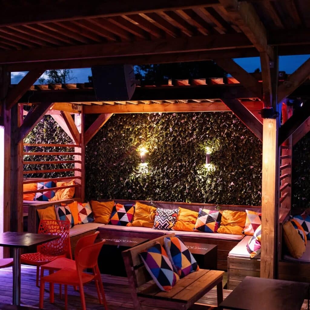 Patio de bar/restaurant sous pergola en bois, avec banquettes, coussins colorés, mur végétal éclairé et sièges. Ambiance conviviale.