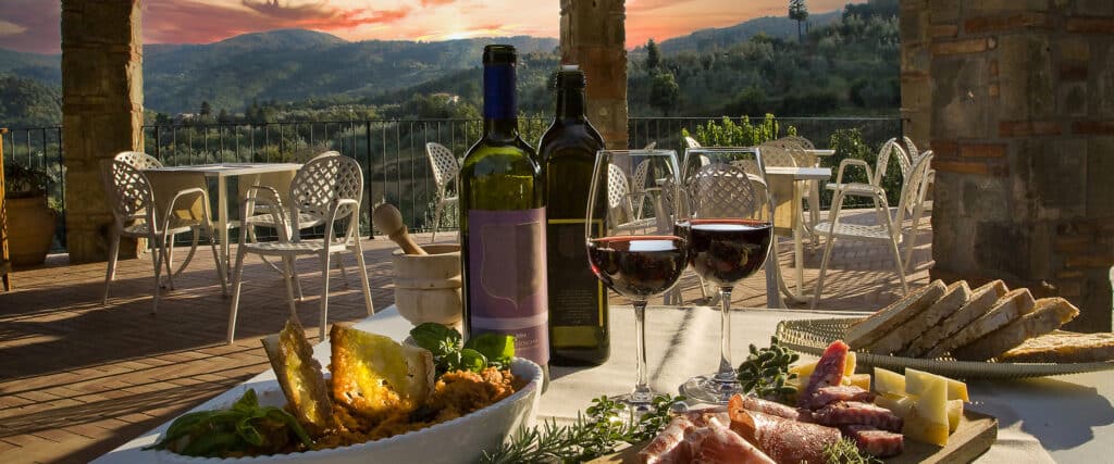 Table sur une terrasse toscane au coucher du soleil, avec vin rouge, charcuterie, fromage, pain, vue sur collines verdoyantes.