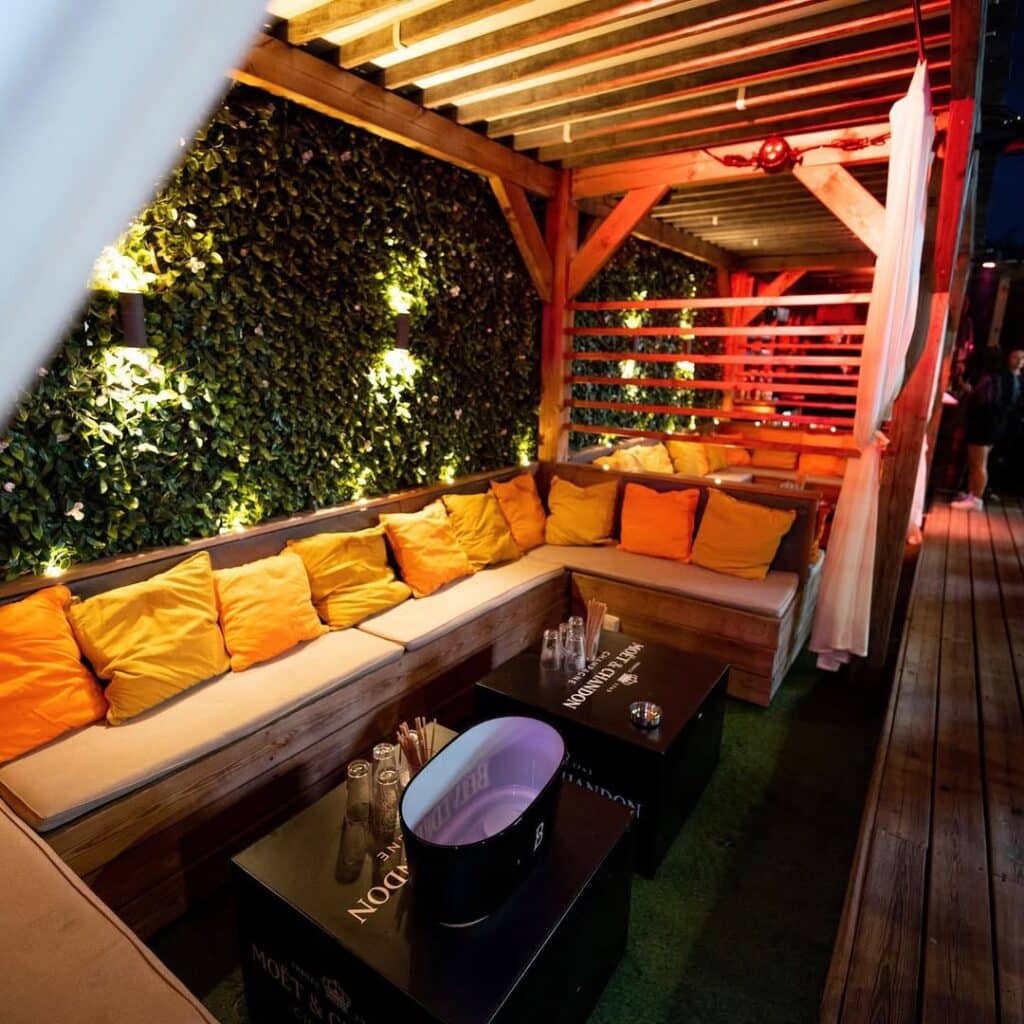 Lounge extérieur chic avec pergola en bois, mur végétal illuminé et coussins jaunes. Tables Moët & Chandon, ambiance festive éclairée en rouge.