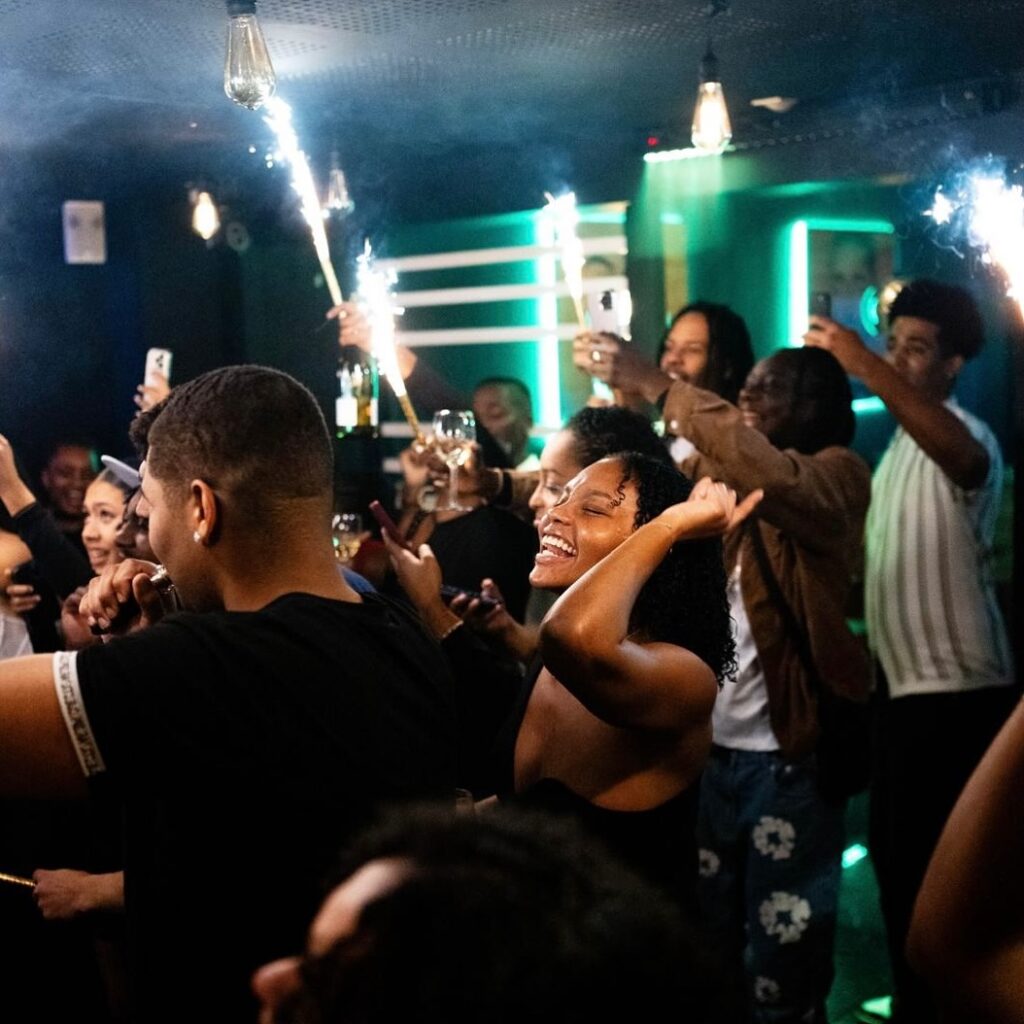 Foule joyeuse célébrant dans un club, étincelles et lumières vertes. Des personnes rient, d'autres filment des moments festifs avec des sparklers.