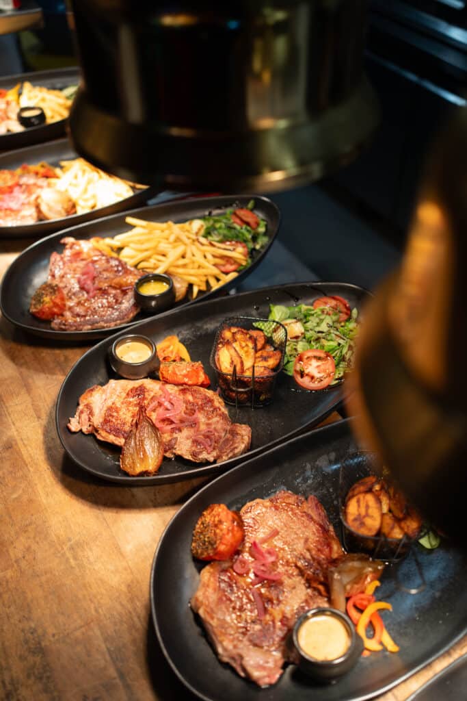 Plats de steaks grillés avec frites, pommes de terre rôties, salades et sauces, alignés sur un comptoir en bois de restaurant.